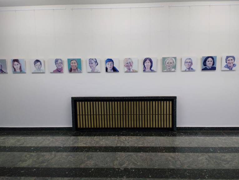 An einer Wand sind dreizehn gemalte Portraits zu sehen. Sie haben alle die gleiche Größe und hängen auf gleicher Höhe.